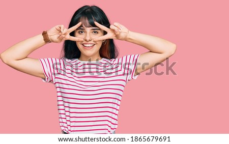Young hispanic girl wearing casual clothes doing peace symbol with fingers over face, smiling cheerful showing victory 
