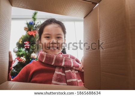 merry christmas and happy holidays. cheerful cute child girl opening a christmas present. little kid having fun near christmas tree indoors. view from inside of the box.