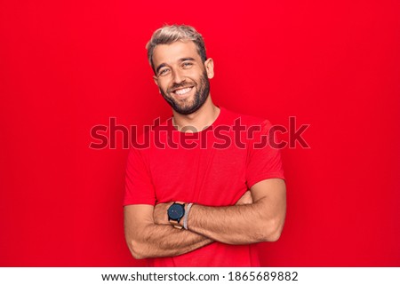 Young handsome blond man wearing casual red t-shirt standing over isolated red background happy face smiling with crossed arms looking at the camera. Positive person.
