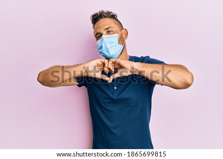 Handsome man with beard wearing medical mask smiling in love doing heart symbol shape with hands. romantic concept. 