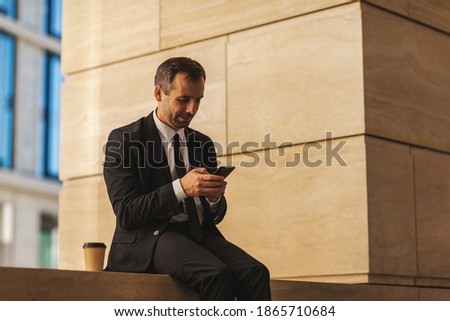 Middle aged businessman in suit sitting in street browsing Internet on cell phone at break, takeaway coffee cup nearby