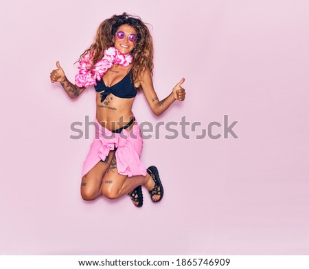 Young beautiful curly woman with tattoo on vacation wearing bikini, sunglasses and hawaiian lei smiling happy. Jumping with smile on face doing ok sign with thumbs up over isolated pink background.