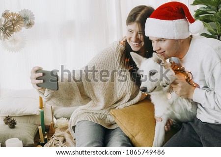 Stylish couple with puppy in lights and santa hat celebrating christmas holidays with family online. Happy young family with cute dog in video call or taking selfie on phone in room with decor