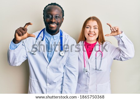 Young interracial couple wearing doctor uniform and stethoscope smiling and confident gesturing with hand doing small size sign with fingers looking and the camera. measure concept. 