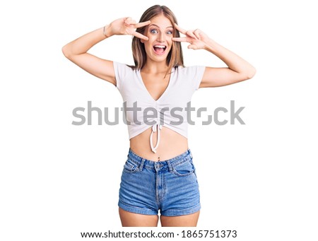 Young beautiful blonde woman wearing casual white tshirt doing peace symbol with fingers over face, smiling cheerful showing victory 