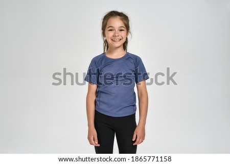Little sportive girl child in sportswear smiling at camera, while standing isolated over white background. Sport, training, fitness, active lifestyle concept. Front view. Horizontal shot