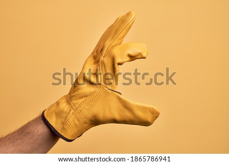 Hand of caucasian young man with gardener glove over isolated yellow background picking and taking invisible thing, holding object with fingers showing space