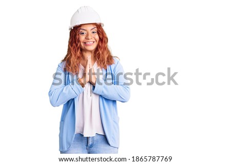 Young latin woman wearing architect hardhat praying with hands together asking for forgiveness smiling confident. 