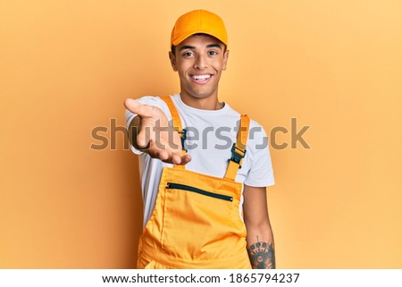 Young handsome african american man wearing handyman uniform over yellow background smiling cheerful offering palm hand giving assistance and acceptance. 