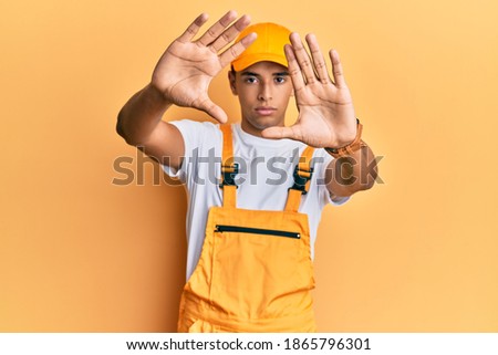 Young handsome african american man wearing handyman uniform over yellow background doing frame using hands palms and fingers, camera perspective 