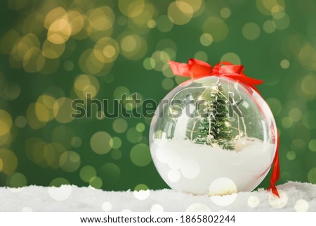 Beautiful transparent Christmas ornament with small fir tree and fairy lights on snow against green background, bokeh effect. Space for text
