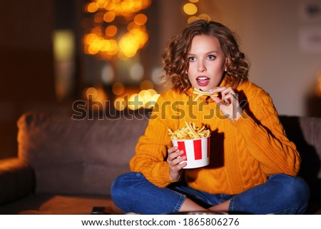 Fascinated young female in sweater eating French fries and watching interesting film on TV while sitting on couch in evening at home
 Royalty-Free Stock Photo #1865806276