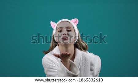 A woman in a white robe, a mask of carbonated bubble clay on her face, blows a kiss