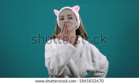 A woman in a white robe, a mask of carbonated bubble clay on her face, blows a kiss