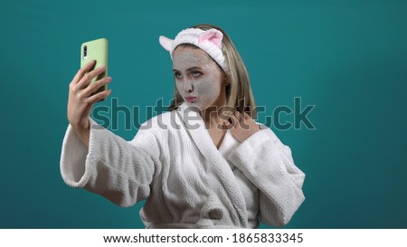 A woman in a white robe, a mask of carbonated bubble clay on her face, takes a selfie