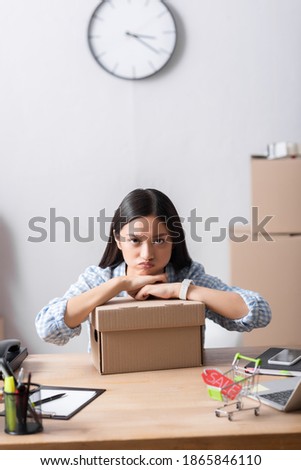 Offended asian volunteer looking at camera while leaning on carton box on desk in charity center on blurred background