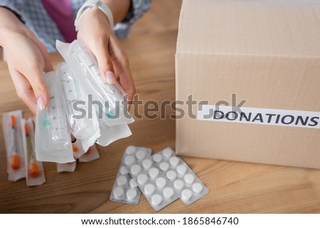Cropped view of syringes in hands of volunteer near pills and box with donations lettering 