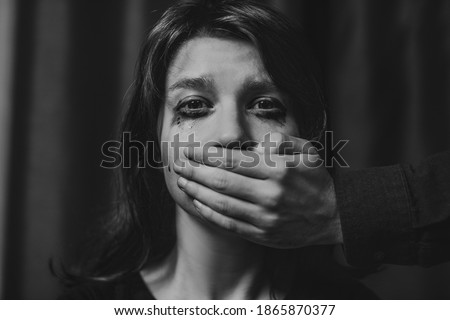 Portrait of a young suffering sad woman with tears in her eyes, a man closes her mouth with his hand. Domestic violence, crying, religion, disagreement, fight, divorce, beating a weaker person, dark. Royalty-Free Stock Photo #1865870377