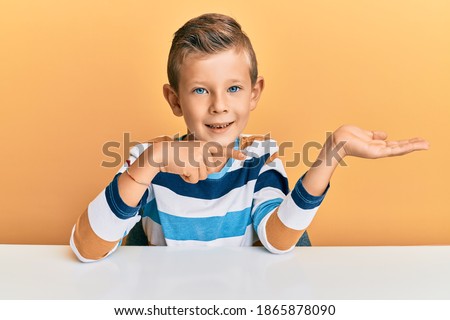 Adorable caucasian kid wearing casual clothes sitting on the table amazed and smiling to the camera while presenting with hand and pointing with finger. 