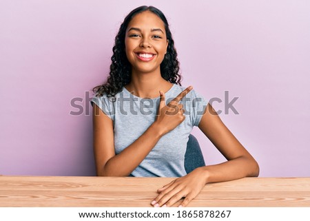 Young african american girl wearing casual clothes sitting on the table cheerful with a smile of face pointing with hand and finger up to the side with happy and natural expression on face 