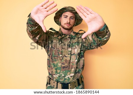 Young caucasian man wearing camouflage army uniform doing frame using hands palms and fingers, camera perspective 