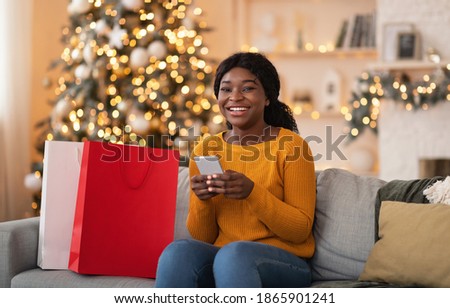 Online purchases and reviews of stores, cashback for buyers. Happy millennial african american woman with smartphone sit on sofa with paper bags with purchases in cozy interior of home with garlands