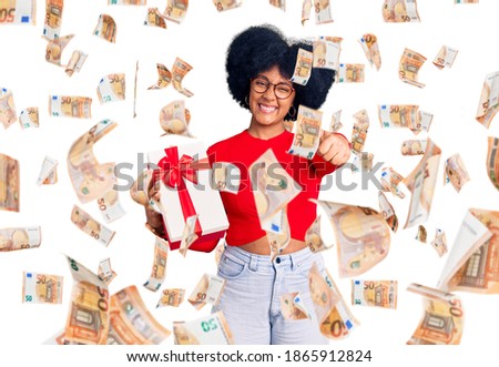 Young african american girl holding gift approving doing positive gesture with hand, thumbs up smiling and happy for success. winner gesture.