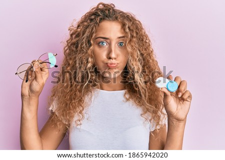Beautiful caucasian teenager girl holding glasses and contact lenses puffing cheeks with funny face. mouth inflated with air, catching air.  Royalty-Free Stock Photo #1865942020