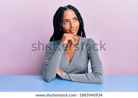 Beautiful hispanic woman wearing casual clothes sitting on the table with hand on chin thinking about question, pensive expression. smiling with thoughtful face. doubt concept. 