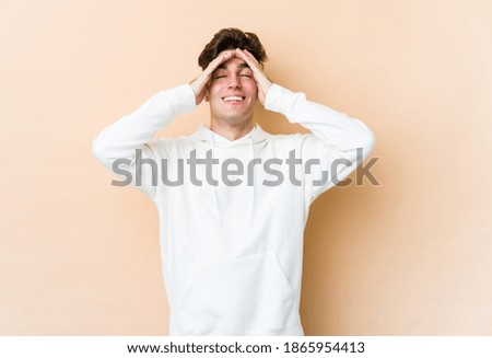 Young caucasian man isolated on beige background laughs joyfully keeping hands on head. Happiness concept.