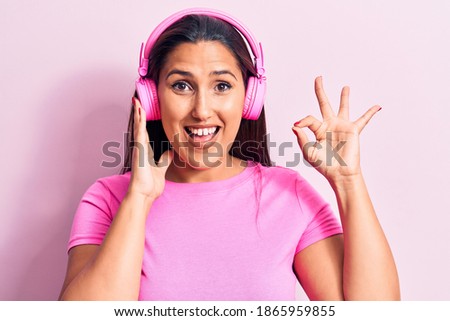 Young beautiful brunette woman listening to music using headphones doing ok sign with fingers, smiling friendly gesturing excellent symbol 