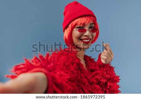 Wonderful short haired girl with bright lips and trendy sunglasses in red fashionable clothes smiling and making selfie..