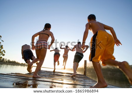 Rear ground view of six family members in swimwear jumping off a jetty into the lake at sunset.