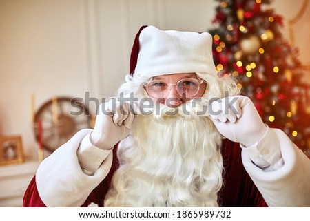 Portrait of Santa Claus in glasses touching his beard near the fireplace and christmas tree with gifts. New year and Merry Christmas , happy holidays concept