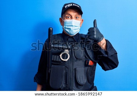 Senior police man wearing surgical mask smiling happy and positive, thumb up doing excellent and approval sign 