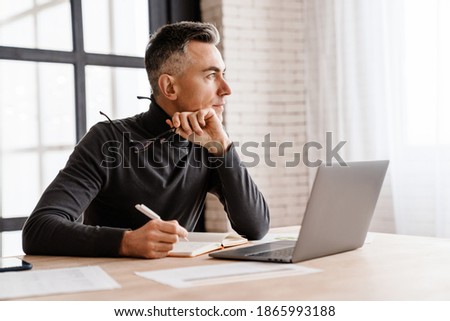 Handsome serious man working with laptop and planner while sitting at table indoors
