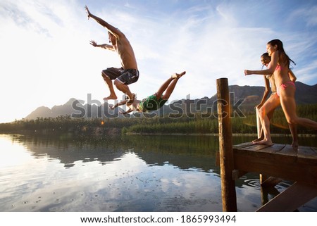 Horizontal shot of two guys diving off the jetty into a lake with two girls preparing to follow them.