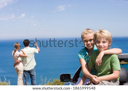 Horizontal shot of a brother and sister sitting on a car's bonnet overlooking the ocean with their parents taking self portrait in the background.