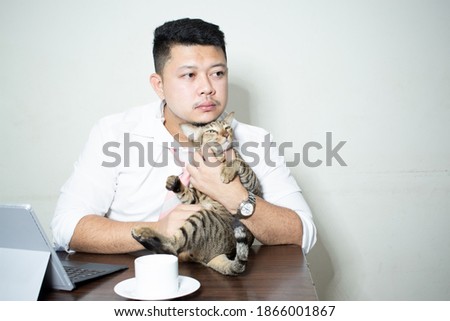 Young bearded businessman sits in desk and uses laptop, hugging a cute cat. In a white background. Man working, blogging. On table is  cup of coffee.