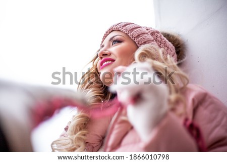 Pretty blond woman dressed in pink warm clothes, padded jacket, white sweater, pants and knitted hat with fur pompon and holding winter fuchsia lace-up skates for figure skating posing in the street 