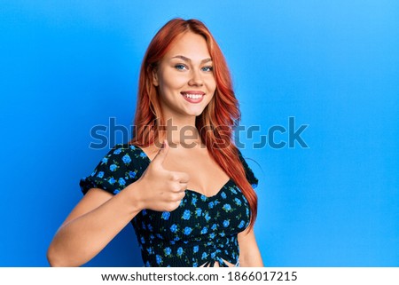 Young beautiful redhead woman wearing casual clothes over blue background doing happy thumbs up gesture with hand. approving expression looking at the camera showing success. 