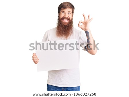 Handsome young red head man with long beard holding blank empty banner doing ok sign with fingers, smiling friendly gesturing excellent symbol 