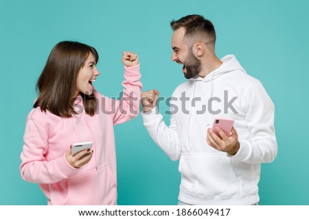 Happy young couple two friends man woman 20s wearing white pink casual hoodie using mobile cell phone typing sms message doing winner gesture isolated on blue turquoise background studio portrait