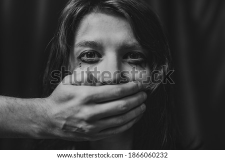 Portrait of a young suffering sad woman with tears in her eyes, a man closes her mouth with his hand. Domestic violence, crying, religion, disagreement, fight, divorce, beating a weaker person, dark. Royalty-Free Stock Photo #1866060232