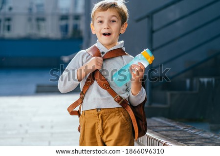 Cute blond little boy going first time to school. Pupil of primary school holding bottle with backpack at sunny day outdoor Walking on School Campus.. Royalty-Free Stock Photo #1866096310