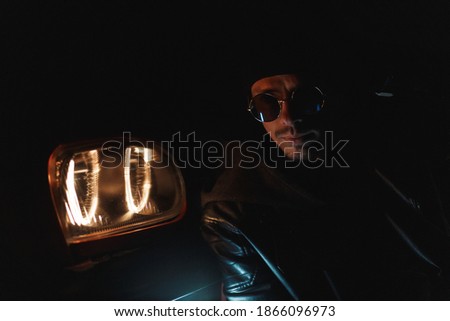 fashionable man with stylish sunglasses in black leather jacket sits near headlights at night