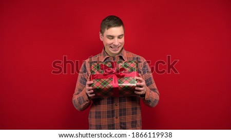 A young man shakes a Christmas present in his hands and rejoices. Shooting in the studio on a red background.