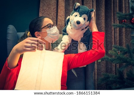 Young teenage girl in a red dress with a soft toy dog sitting at home on Christmas, New Year's eve with medical mask, waiting for guests