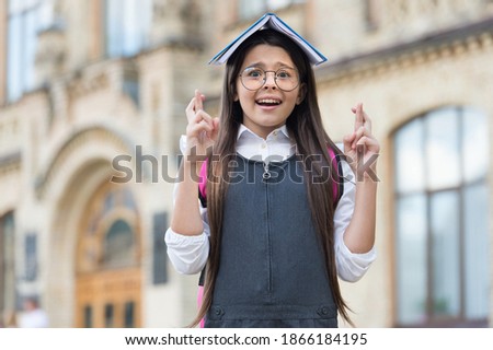 Superstitious little kid in uniform hold school book on head keeping fingers crossed outdoors, learning.