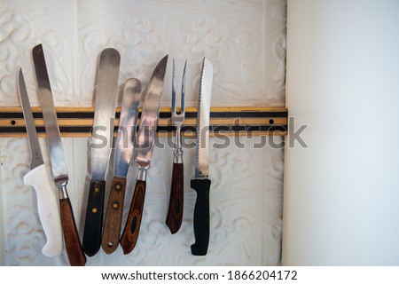 A long black and brass colour strip magnet knife rack holder on a white wall. The holder has a number of long stainless steel sharp knives, icing spatulas and a meat fork with black plastic handles. 